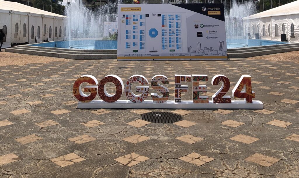 Outdoor setup for the Global Off-Grid Solar Forum and Expo 2024 (GOGSFE24) at the Kenyatta International Convention Centre in Nairobi, Kenya. In the foreground, colorful letters spell out ‘GOGSFE24’ with traditional African patterns. Behind, a program board, fountain, and the iconic cylindrical tower of the convention center rise against a partly cloudy sky.